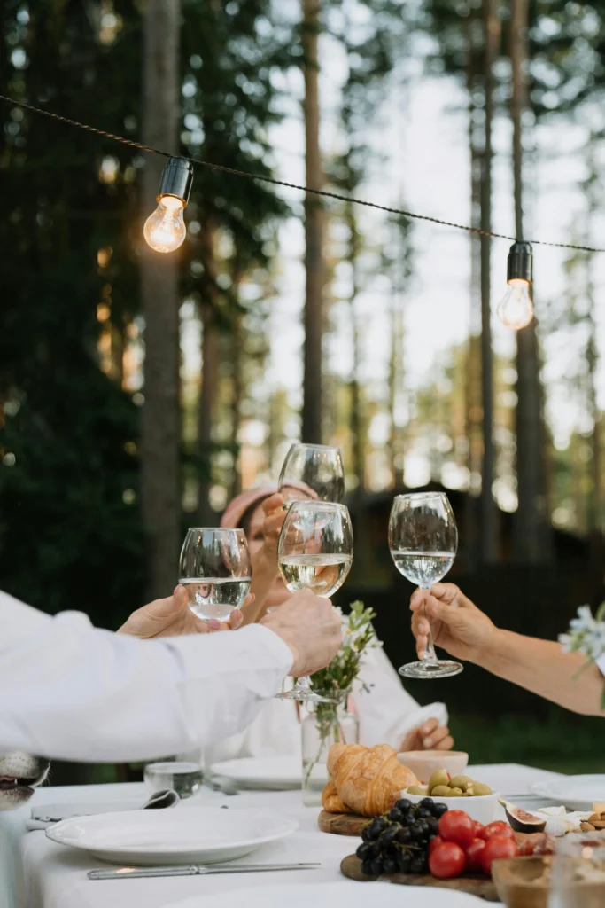 Persone che brindano con bicchieri di vino bianco durante una cena all'aperto, circondati dalla natura e da luci decorative.